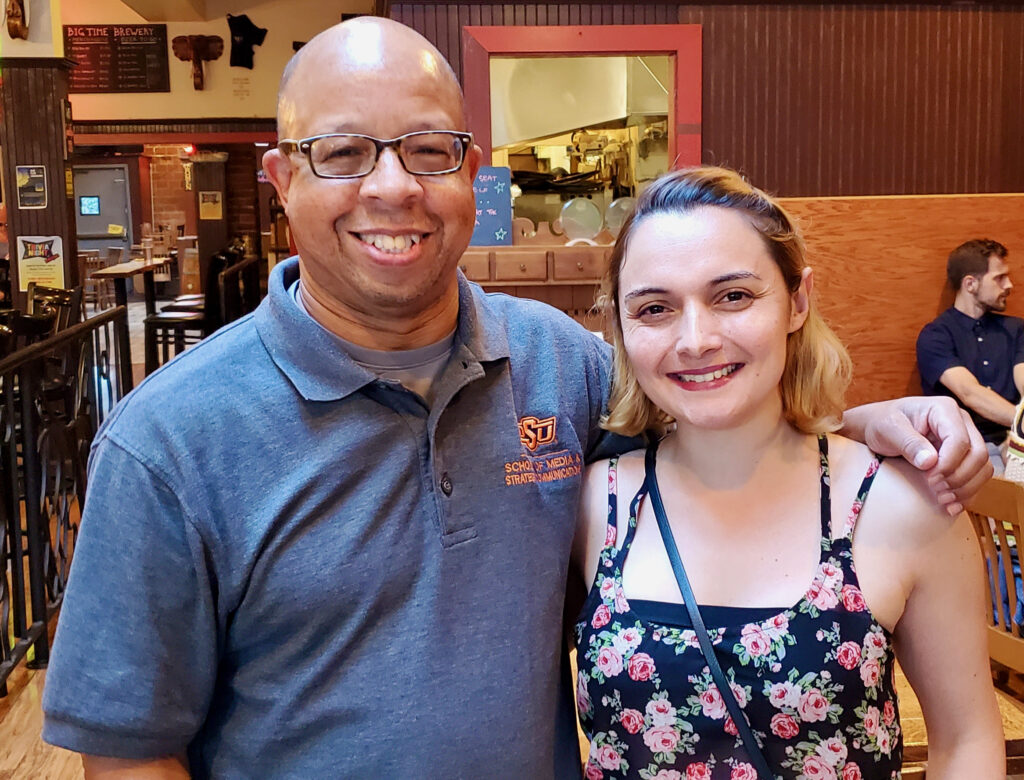 Two adults posing side by side together and smiling. One has their arm around the other.
