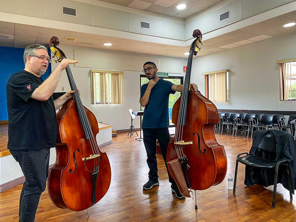 Jamie Ousley, standing with a bass instrument, talks to a student, also with a bass instrument, inside of a music classroom.