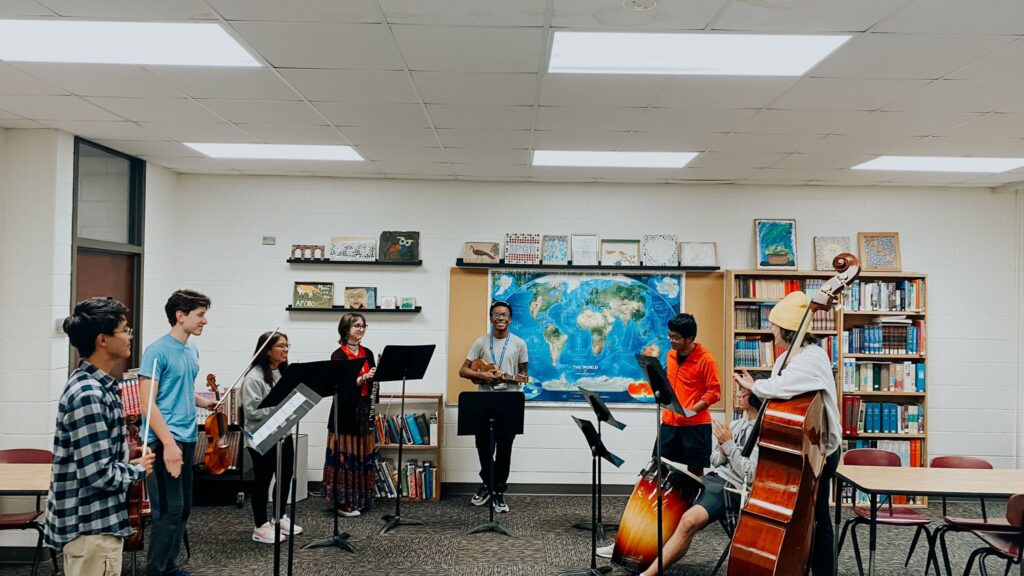 World Learning’s Youth Ambassador Brayan Weelkly Prendigan stands in a semi-circle with six other high school students in a classroom. A map and books line the walls, and all the students are holding different musical instruments.