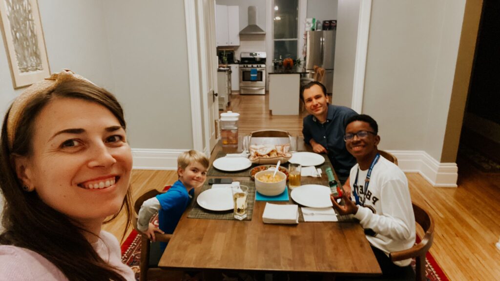 World Learning’s Youth Ambassador Brayan Weelkly Prendigan is sitting with his host family at a square dining room table. He is next to the father, a young boy is across from him, and a mother is taking the selfie shot.