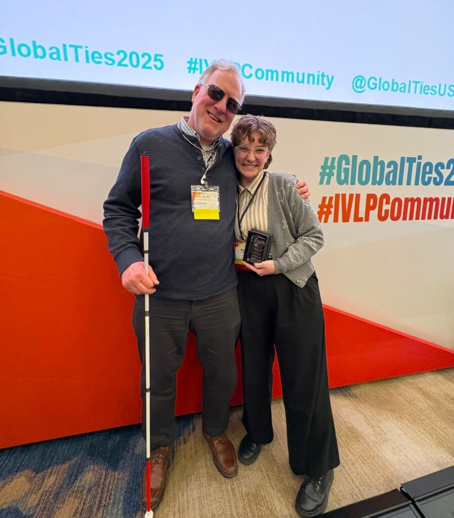 Two World Learning staff members (a man and a woman) stand side by side in front of a large backdrop in red and white. The man holds a walking cane, and the woman holds an award plaque.