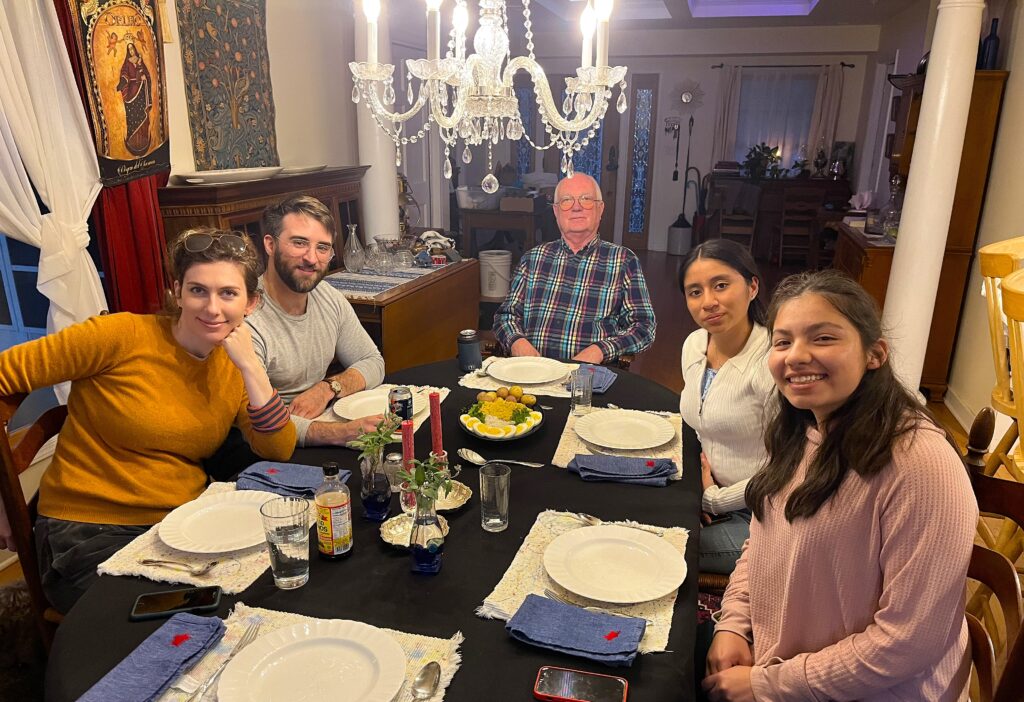 Two Youth Ambassadors female high school students sit with three adults around a dining room table before dinner.