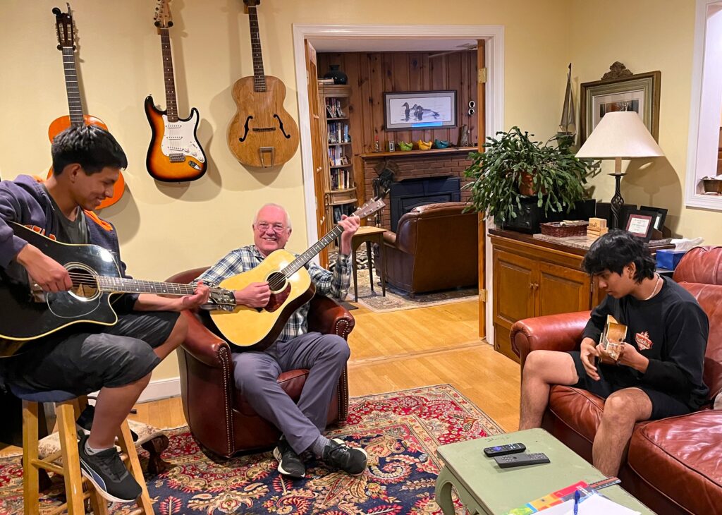 Two Youth Ambassadors male students playing guitar with an older man. They are sitting in a living room.