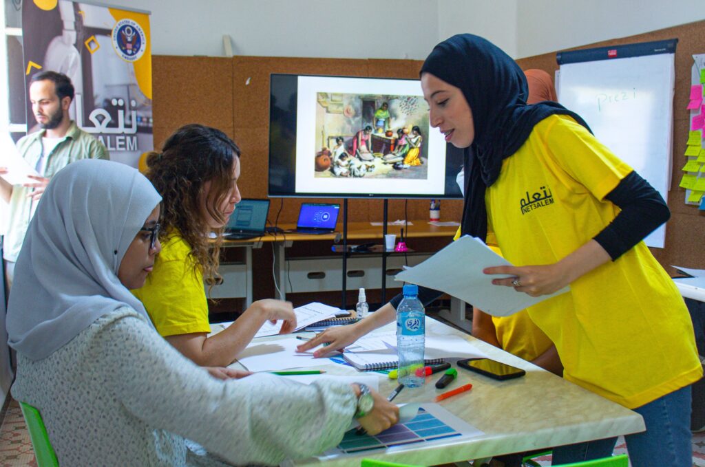 Two women are sitting at a small table covered with papers, markers, post-it notes, a calculator, and a water bottle. A third woman is holding a stack of papers and is leaning over the table, pointing at something on the table, helping the two sitting.