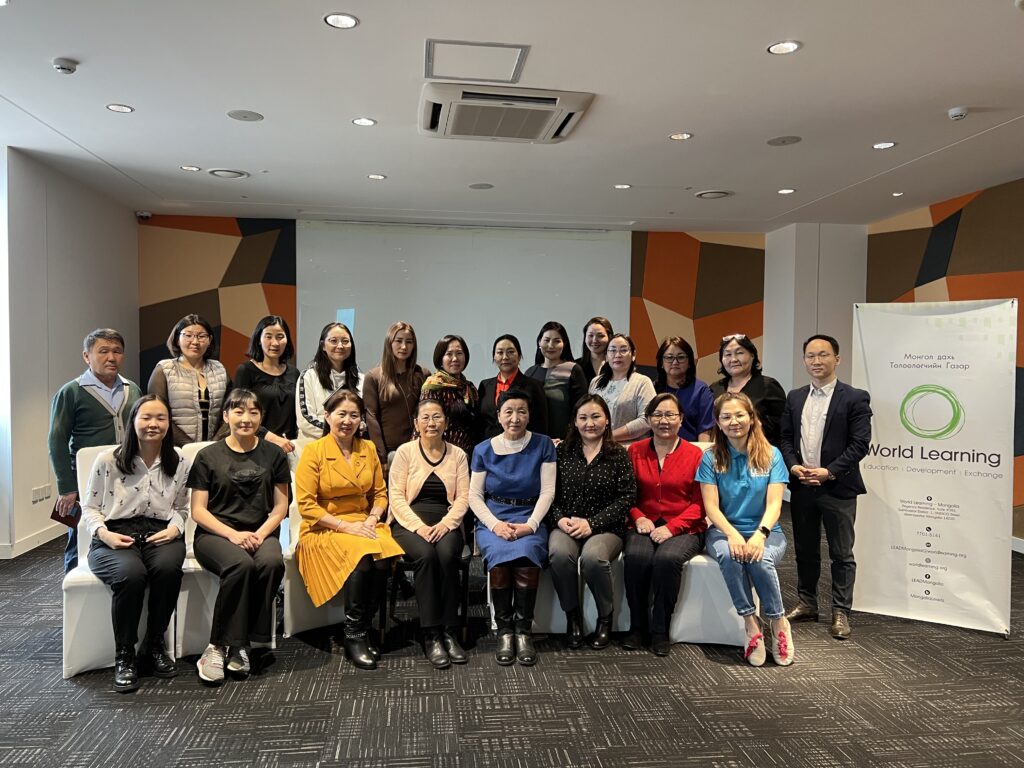 21 adults pose for a group photo with half standing and half sitting next to a banner that contains the World Learning logo.