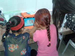 Two young girls work at a small orange computer as an adult woman looks on.