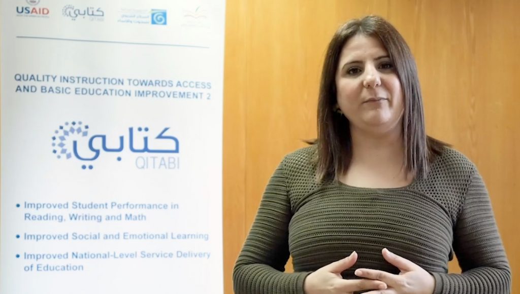 A woman in a green sweater stands facing the camera with her hands clasped in front of her. A banner in Arabic and Englishis behind her to the left.