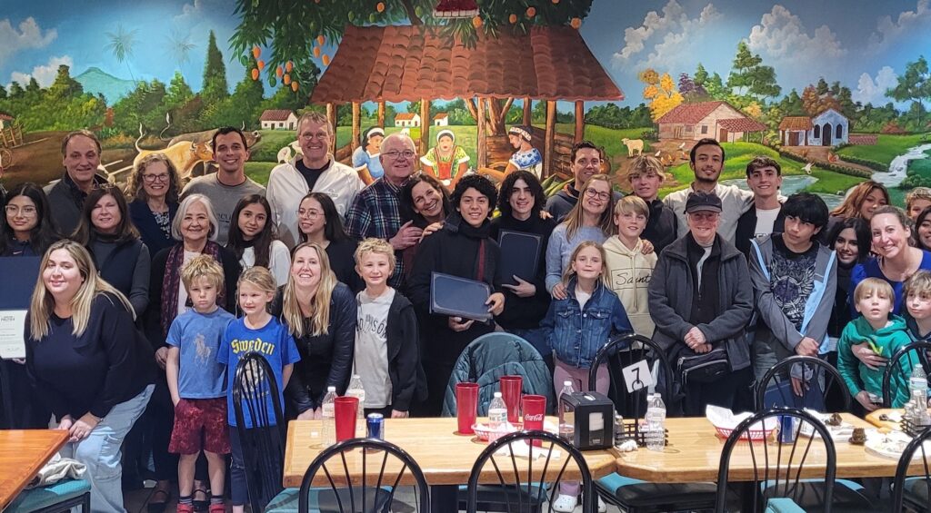 A large group of Youth Ambassador students and family members are gathered in front of table, posing for a photo. A mural is on the wall behind the group of a countryside scene.
