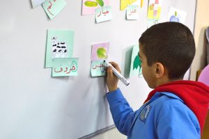 A young boy stands at a white board and writes in Arabic.