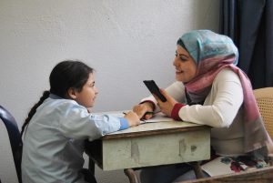 A woman and a young girl sit at a table across from one another. The woman holds the girl's hand reassuringly.