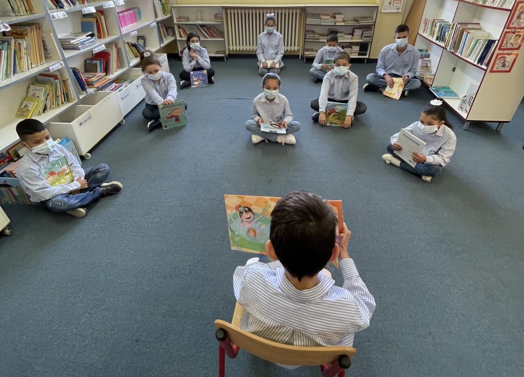 A boy sits in front of his class reading a book to a group of children wearing masks.