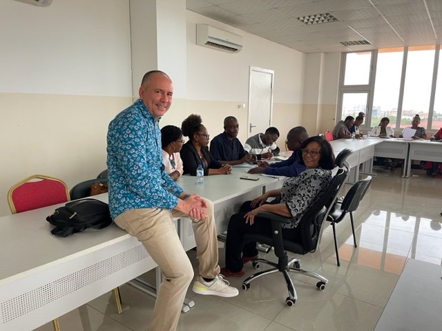 A room of adults gathered in chairs around tables, with one sitting on a table looking at the camera.
