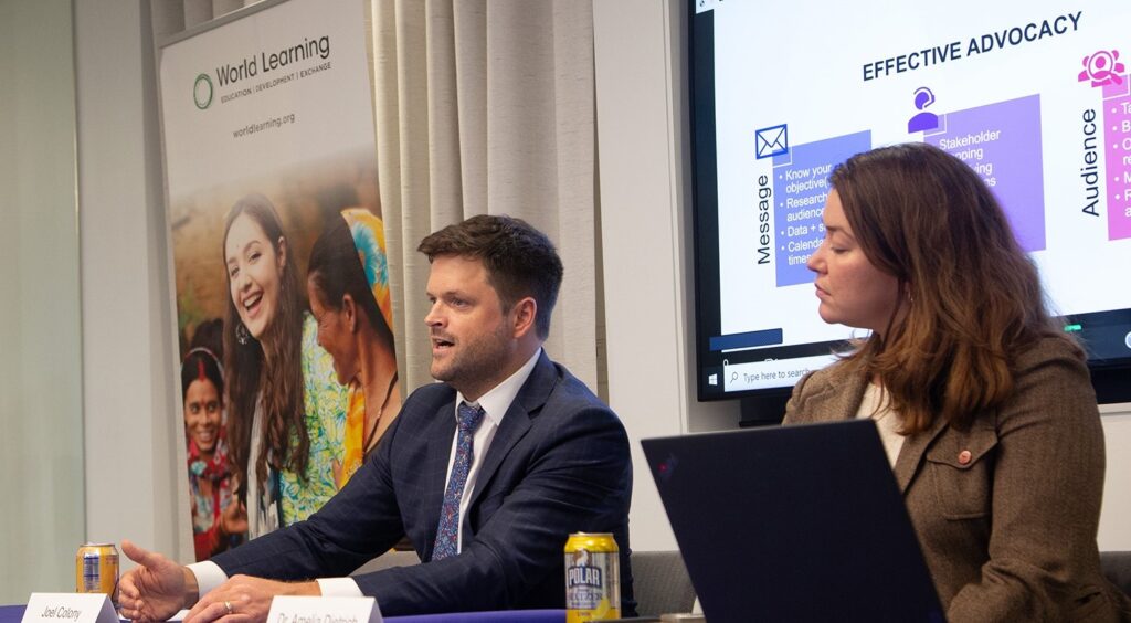 World Learning’s Vice President for External Engagement and Advocacy Joel Colony sits at a table next to a woman. Behind them is a World Learning banner and a projection of a presentation.
