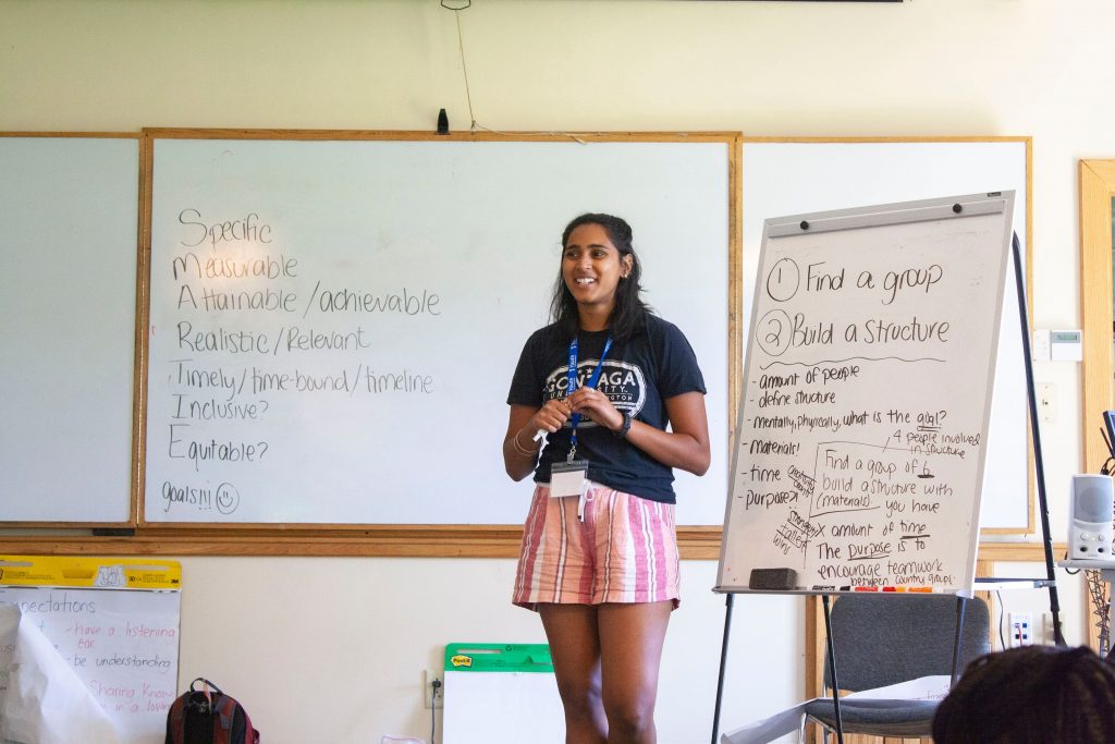 A woman stands in front of a white board with a flipchart next to her.
