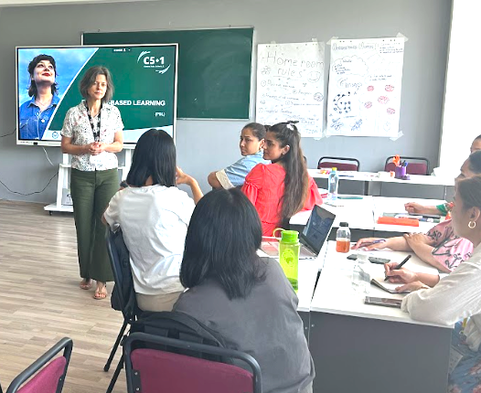 World Learning’s Dr. Kara McBride stands in the front of a classroom and listens to a group of adult teachers who sit at tables. A large screen with a presentation is set up behind her.