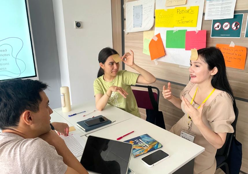 Three adults sit at a table talking with post it notes on their foreheads during World Learning’s C5 + O.N.E. Ed program.