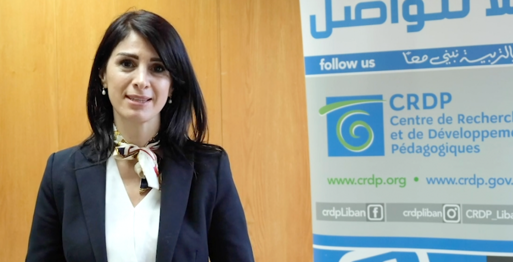 A woman in a dark blue blazer with a white shirt and scarf around her neck stands facing the camera. A banner in Arabic and French is behind her to the right.