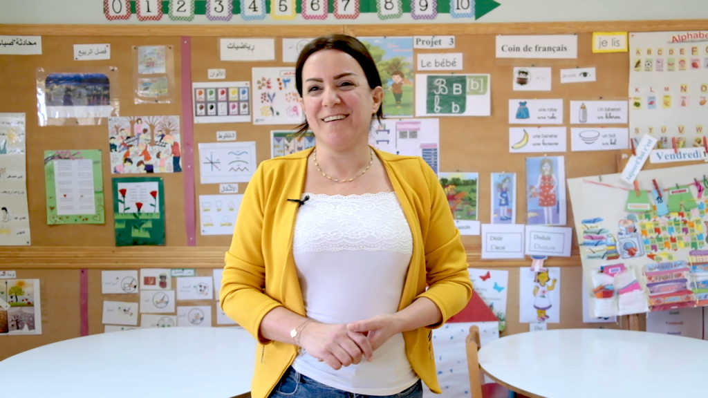 A woman in a white shirt and yellow cardigan stands looking at the camera in a classroom.