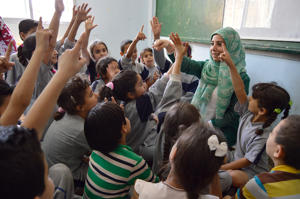 A woman stands in front of her class surrounded by young students.